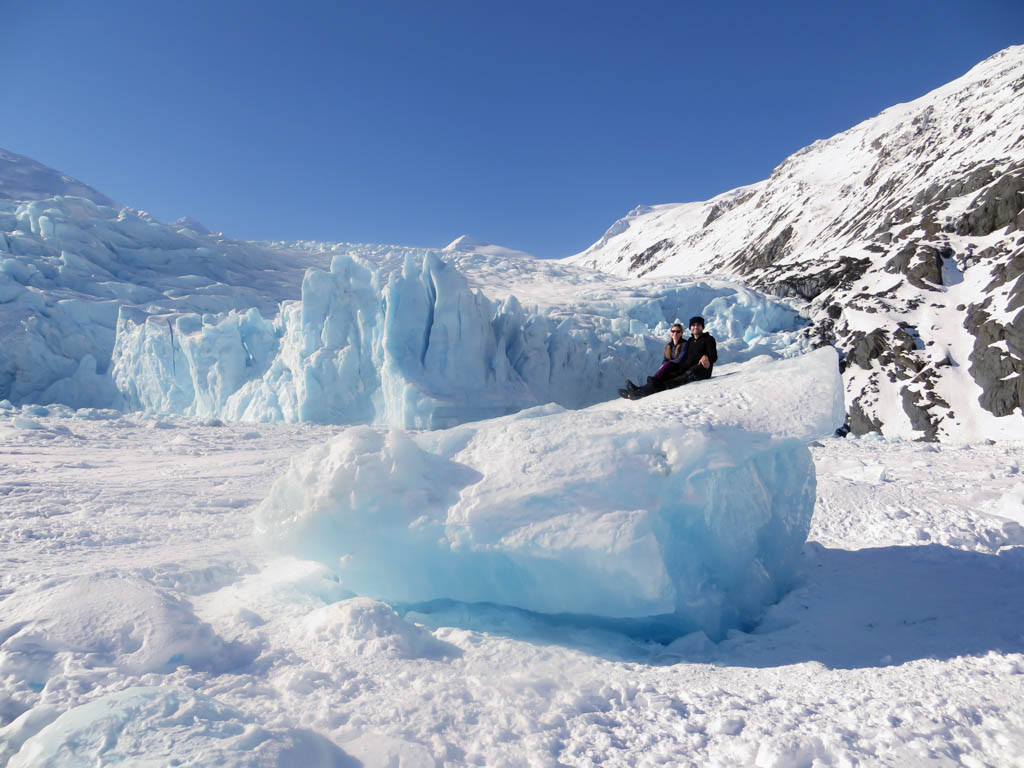 Two people sitting on top of an iceberg frozen in Portage lake with the Portage Glacier in the background. The shear size of the glacier & iceberg make the people look so tiny.