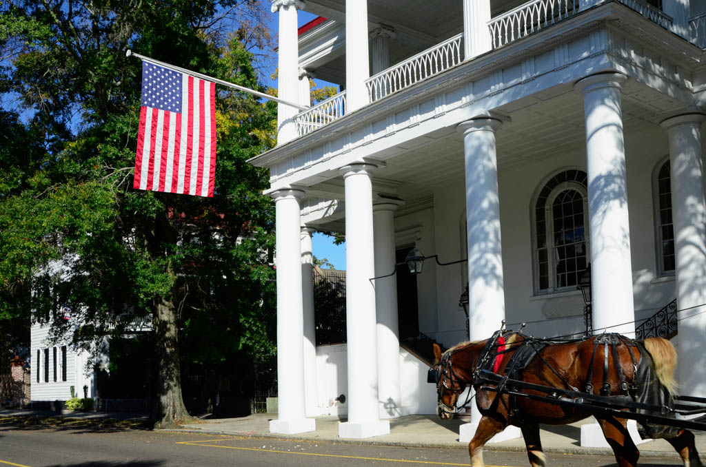 A horse-drawn carriage makes its way past a historical building with an American flag hanging from the portico.