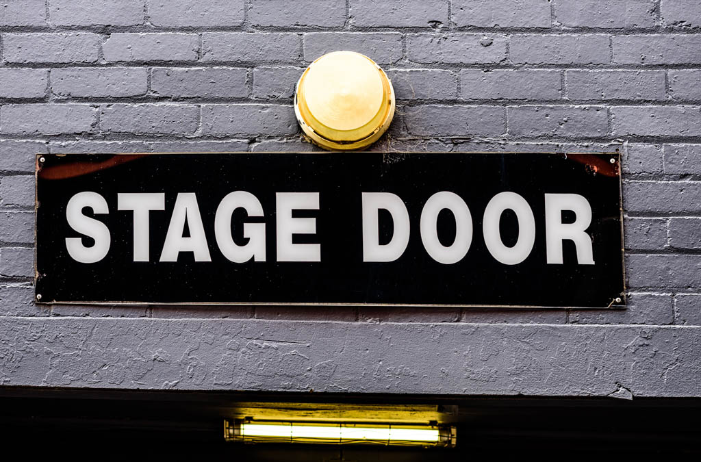 A diagonal 'Stage Door' sign with white on black lettering, on a brick wall and a lamp above