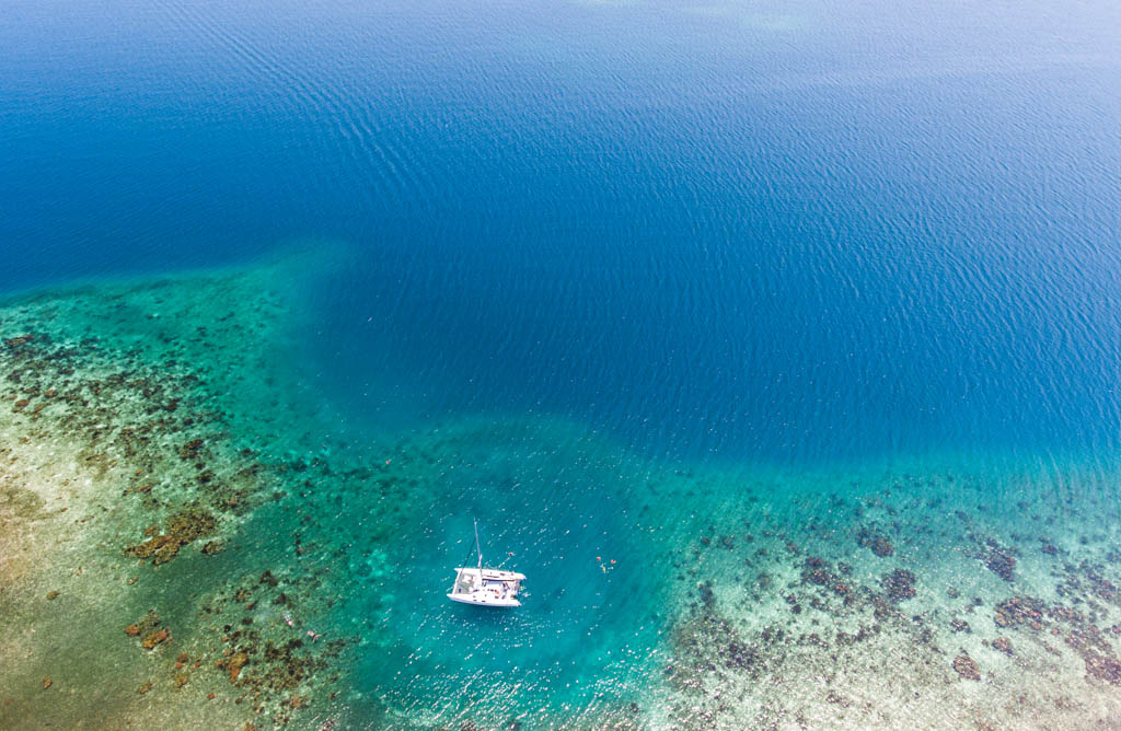 An aerial image of a catamaran anchored near a tropical coral reef.