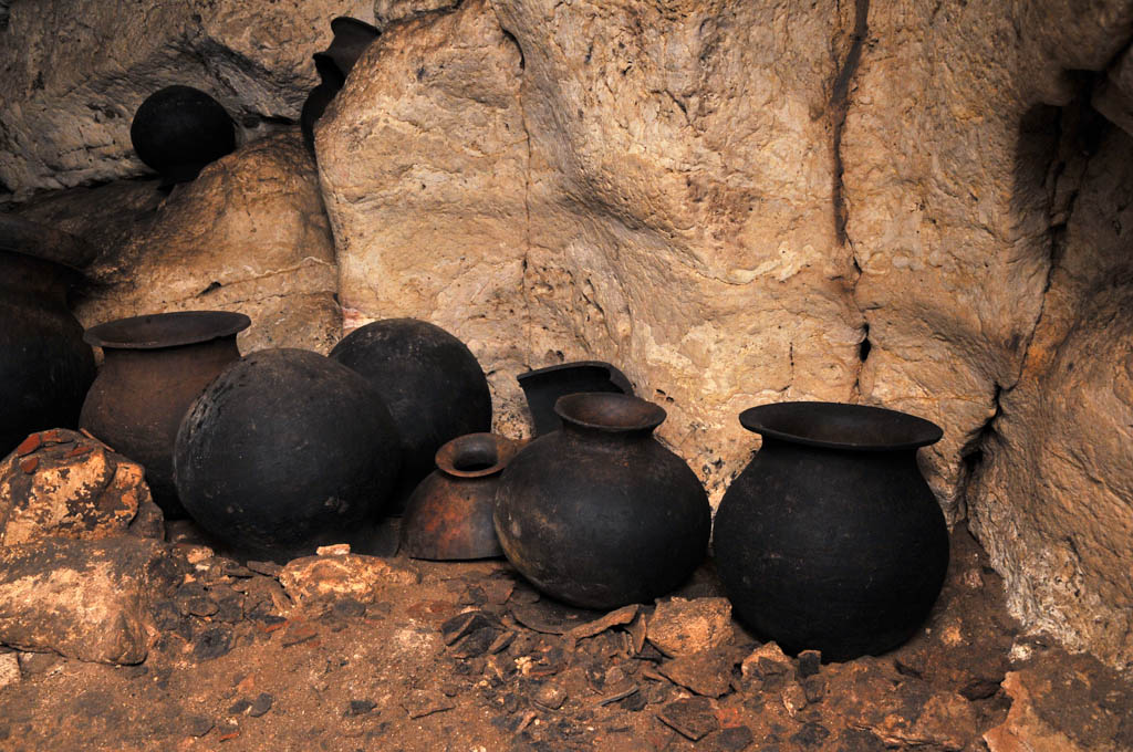 Several ancient Mayan clay pots in a row along the wall of a natural limestone cave. Pot shards litter the floor.