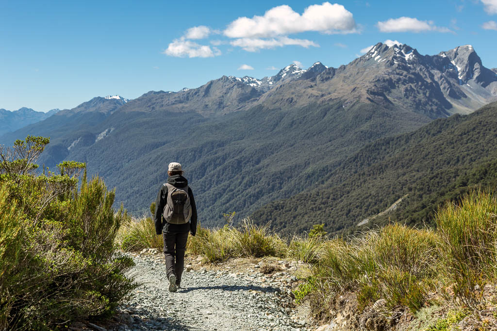 DSLR picture of a woman hiker with a backpack on footpath at Key Summit Lookout Trail, New Zealand. The woman is alone and she is walking in the direction of the mountain in front. The sky is blue and the mountains are visible in the background.