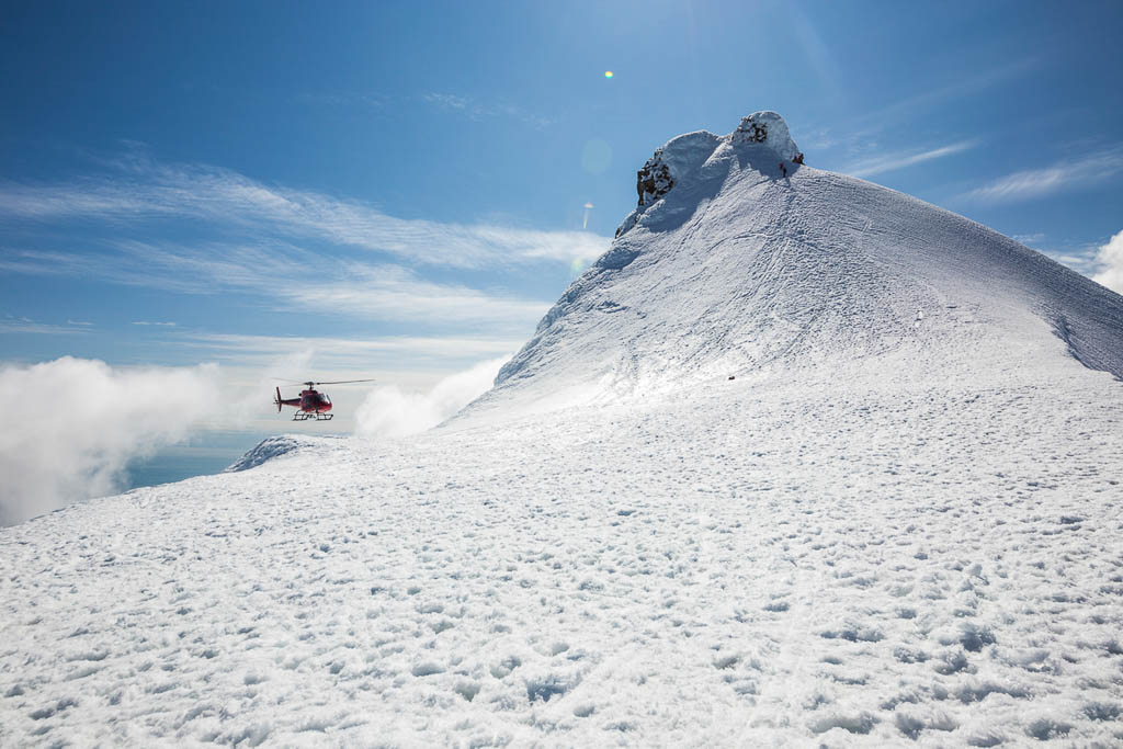The picture shows the snow-capped peak of a mountain, on which a helicopter is landing and a closer look reveals two climbers.