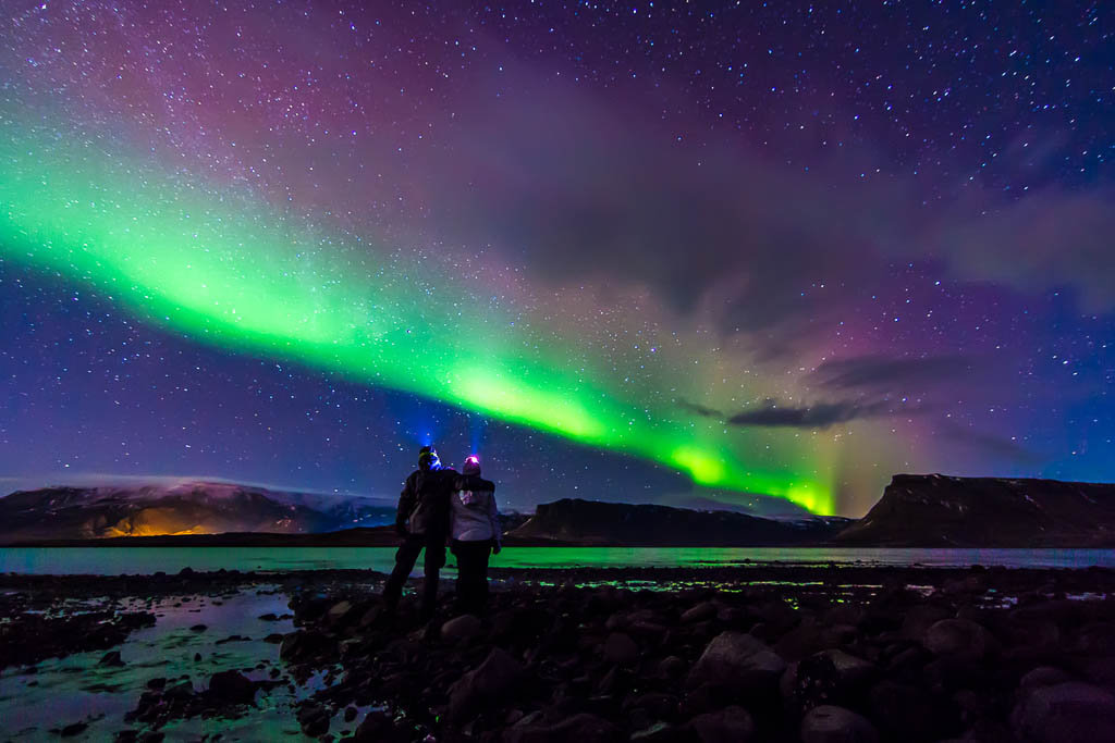 Young adventurist couple hugged while gazing in the dark night sky under the spectacular celestial lights Aurora Borealis, which makes Iceland popular spot for tourist willing to witness one of the greatest natural phenomenon. Shot with Canon EOS, wide angle lens, f2.8, long exposure.