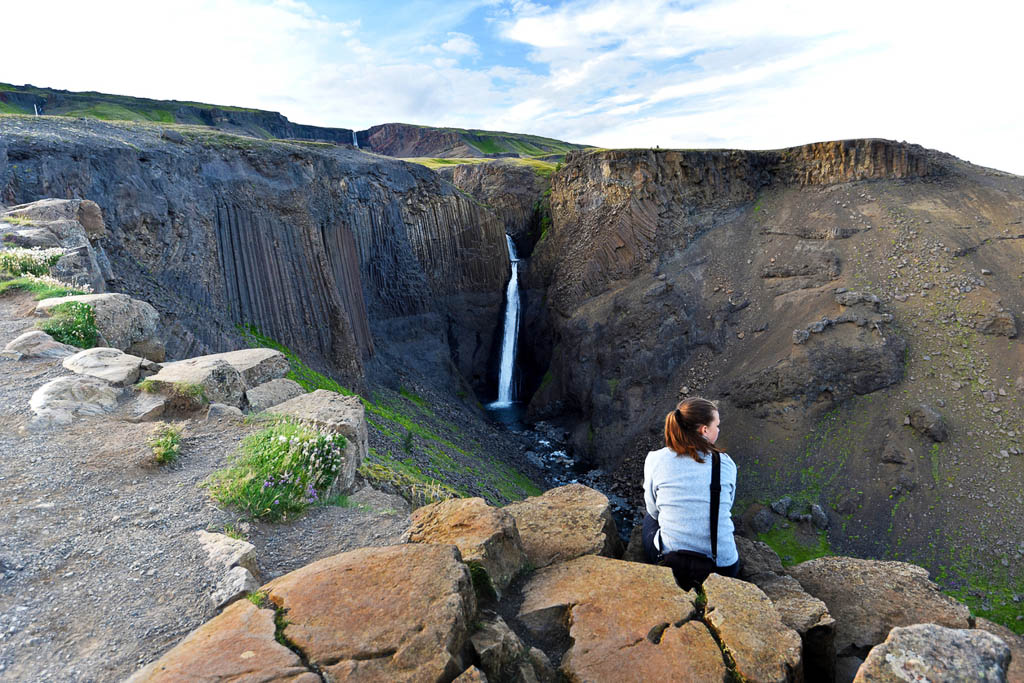 Young woman is sitting at the rock in front of spectacular view at Litlanesfoss waterfall surrounded with vertical basalt columns, Fljotsdalshreppur municipality of Eastern Iceland
