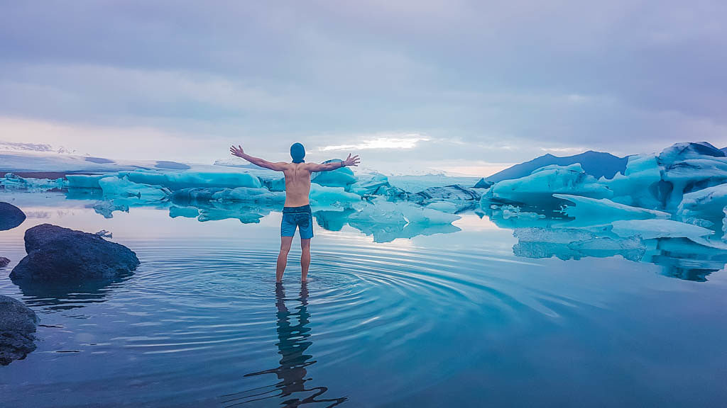 Young man stands in the Glacier lagoon with his arms wide spread. Man wearing only swimming shorts. Ice bergs drifting in the lagoon. Cold temperatures for ice swimming. Calm surface of the water.